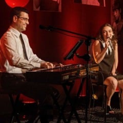Bia Peroni canta sentada ao lado de João Carlos de Luca, que toca teclado sorrindo, em um palco iluminado em vermelho.