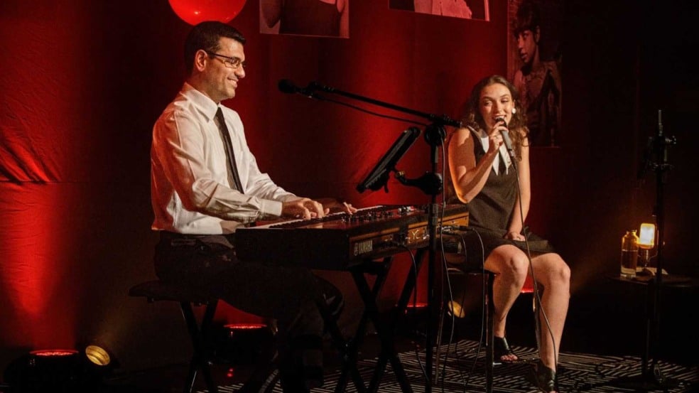 Bia Peroni canta sentada ao lado de João Carlos de Luca, que toca teclado sorrindo, em um palco iluminado em vermelho.