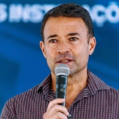 Homem segurando um microfone enquanto fala em um evento, diante de um fundo azul desfocado.