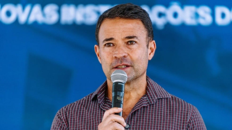 Homem segurando um microfone enquanto fala em um evento, diante de um fundo azul desfocado.