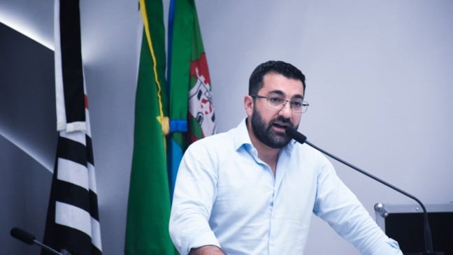 Vereador Faouaz Taha discursando na Câmara Municipal de Jundiaí, usando camisa clara e óculos, com bandeiras ao fundo.