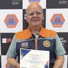 Homem sorrindo, usando colete da Defesa Civil, segurando o Certificado de Resiliência de Jundiaí em frente a um painel com logos da Defesa Civil do Estado de São Paulo.