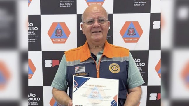Homem sorrindo, usando colete da Defesa Civil, segurando o Certificado de Resiliência de Jundiaí em frente a um painel com logos da Defesa Civil do Estado de São Paulo.