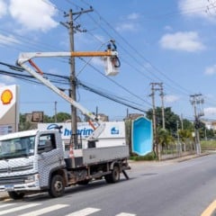 Técnico da Prefeitura de Jundiaí instala luminária LED em poste utilizando cesto elevatório acoplado a caminhão na Rua do Retiro, com postos de combustível e veículos ao fundo.