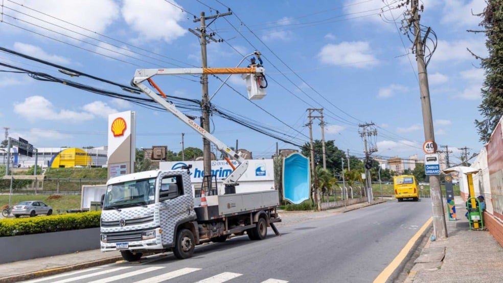 Técnico da Prefeitura de Jundiaí instala luminária LED em poste utilizando cesto elevatório acoplado a caminhão na Rua do Retiro, com postos de combustível e veículos ao fundo. Técnico da Prefeitura de Jundiaí instala luminária LED em poste utilizando cesto elevatório acoplado a caminhão na Rua do Retiro, com postos de combustível e veículos ao fundo.