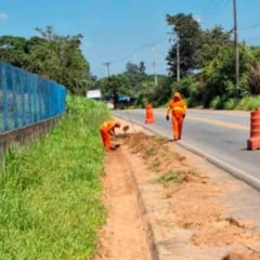 Funcionários da Prefeitura de Jundiaí, com uniformes laranja, limpam e desobstruem canaletas na Avenida da Uva, utilizando ferramentas manuais ao lado de cones de sinalização na pista.