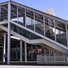 Passarela envidra&ccedil;ada com escada rolante na esta&ccedil;&atilde;o da Linha 7-Rubi, estrutura met&aacute;lica clara e pessoas circulando sob c&eacute;u azul.