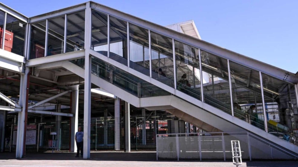 Passarela envidraçada com escada rolante na estação da Linha 7-Rubi, estrutura metálica clara e pessoas circulando sob céu azul.