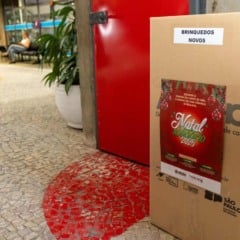 Caixa de arrecadação da Campanha Natal Solidário 2025 com etiqueta “Brinquedos Novos”, posicionada em corredor público, ao lado de um cartaz natalino.