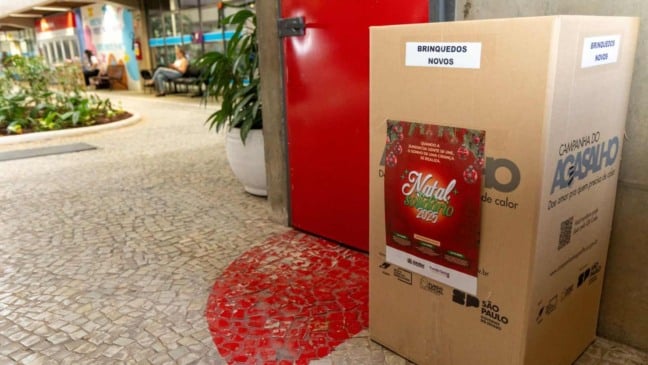 Caixa de arrecadação da Campanha Natal Solidário 2025 com etiqueta “Brinquedos Novos”, posicionada em corredor público, ao lado de um cartaz natalino.