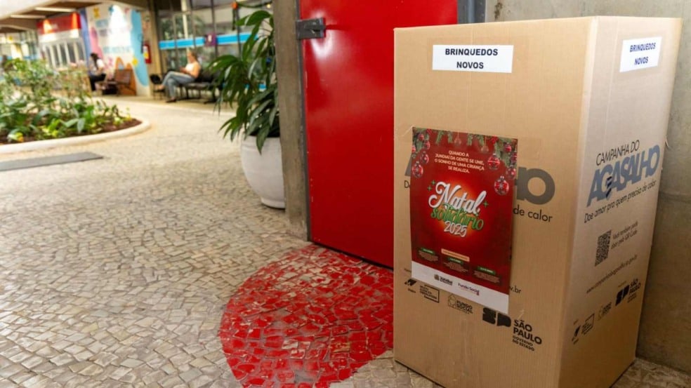 Caixa de arrecadação da Campanha Natal Solidário 2025 com etiqueta “Brinquedos Novos”, posicionada em corredor público, ao lado de um cartaz natalino. Caixa de arrecadação da Campanha Natal Solidário 2025 com etiqueta “Brinquedos Novos”, posicionada em corredor público, ao lado de um cartaz natalino.