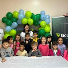 Grupo de crianças e três voluntárias do Sicoob Integrado posam sorrindo em frente a uma decoração de balões verdes e azuis durante uma atividade do Programa Voluntário Transformador.