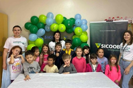 Grupo de crianças e três voluntárias do Sicoob Integrado posam sorrindo em frente a uma decoração de balões verdes e azuis durante uma atividade do Programa Voluntário Transformador.