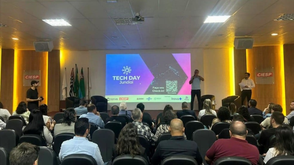 Imagem do auditório do CIESP Jundiaí durante o Tech Day 2025, com público sentado e três apresentadores no palco diante de um telão que mostra o logotipo do evento e um QR Code para check-in. Imagem do auditório do CIESP Jundiaí durante o Tech Day 2025, com público sentado e três apresentadores no palco diante de um telão que mostra o logotipo do evento e um QR Code para check-in.