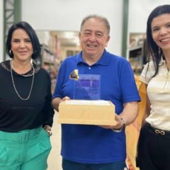 Homem sorridente segura placa comemorativa ao lado de duas mulheres em um dep&oacute;sito da GTS Distribuidora, com caixas de produtos ao fundo.