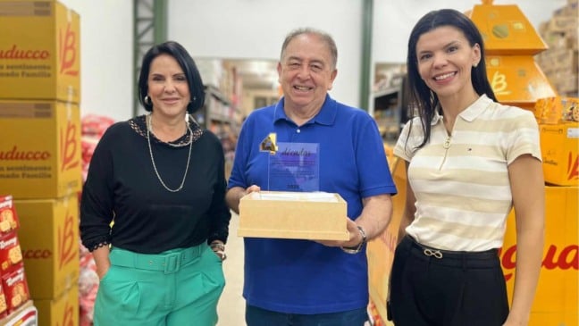 Homem sorridente segura placa comemorativa ao lado de duas mulheres em um depósito da GTS Distribuidora, com caixas de produtos ao fundo.