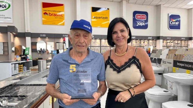 Idoso segura troféu de “5 décadas” ao lado de uma mulher em loja de materiais de construção, durante homenagem da ACE Jundiaí, com expositores e marcas do setor ao fundo.