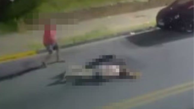 Imagem desfocada mostra uma mulher caída no asfalto durante a noite, enquanto um homem com o rosto pixelado se afasta pela rua.