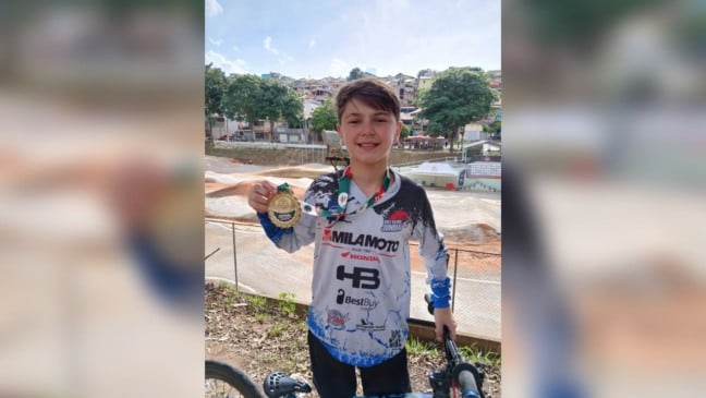 Jovem atleta de BMX Racing de Jundiaí segura medalha dourada e sorri em pista de BMX ao ar livre, ao lado da bicicleta.