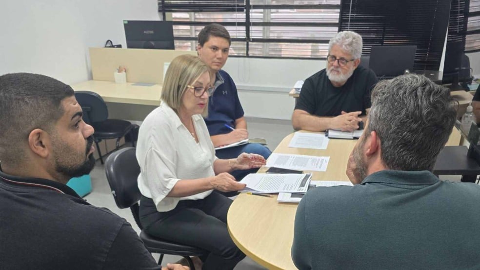 Reunião entre equipe da FMJ e FATEC sentada ao redor de uma mesa, examinando documentos e discutindo detalhes do projeto do aplicativo. Reunião entre equipe da FMJ e FATEC sentada ao redor de uma mesa, examinando documentos e discutindo detalhes do projeto do aplicativo.