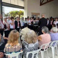 Idosos assistem &agrave; apresenta&ccedil;&atilde;o de um coral em espa&ccedil;o comunit&aacute;rio, com m&uacute;sicos &agrave; frente e moradores sentados em semic&iacute;rculo durante confraterniza&ccedil;&atilde;o.