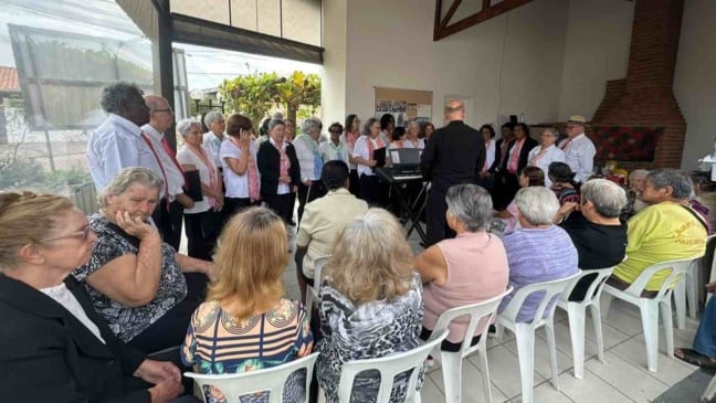 Idosos assistem à apresentação de um coral em espaço comunitário, com músicos à frente e moradores sentados em semicírculo durante confraternização.