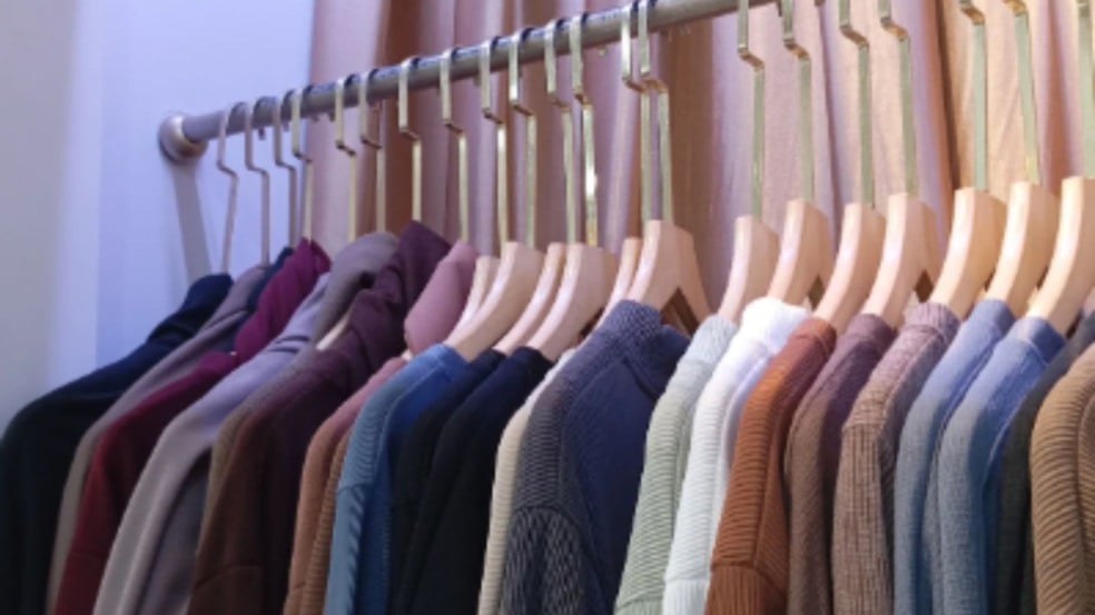 Arara com várias blusas e suéteres em tons neutros e coloridos, pendurados em cabides de madeira, organizados lado a lado.