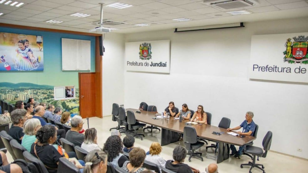 Mesa com quatro representantes da Prefeitura de Jundiaí conduzindo uma reunião com artesãos em um auditório lotado, com placas institucionais da prefeitura ao fundo. Mesa com quatro representantes da Prefeitura de Jundiaí conduzindo uma reunião com artesãos em um auditório lotado, com placas institucionais da prefeitura ao fundo.