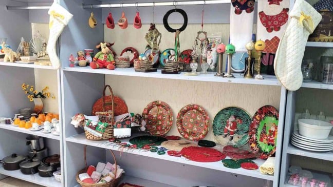 Imagem de prateleiras do Bazar de Natal do Hospital São Vicente, com enfeites natalinos, brinquedos, louças e itens decorativos organizados para venda.