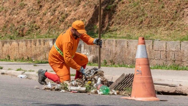 Trabalhador uniformizado limpa boca de lobo em rua de Jundiaí, retirando lixo acumulado ao lado de um cone de sinalização.