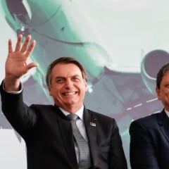 Homem sorrindo e acenando ao lado de outro homem, ambos de terno, em frente a um painel com a imagem de um avião.