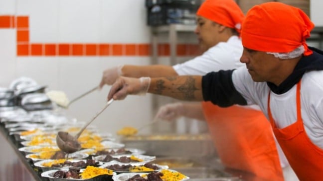 Funcionários do Bom Prato servem refeições quentes em marmitas em cozinha industrial durante o preparo dos almoços.