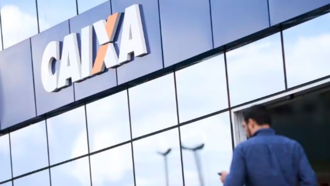 Fachada de agência da Caixa com o logotipo em destaque; homem de costas se aproxima da entrada.