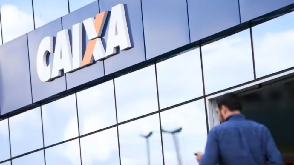 Fachada de agência da Caixa com o logotipo em destaque; homem de costas se aproxima da entrada. Fachada de agência da Caixa com o logotipo em destaque; homem de costas se aproxima da entrada.