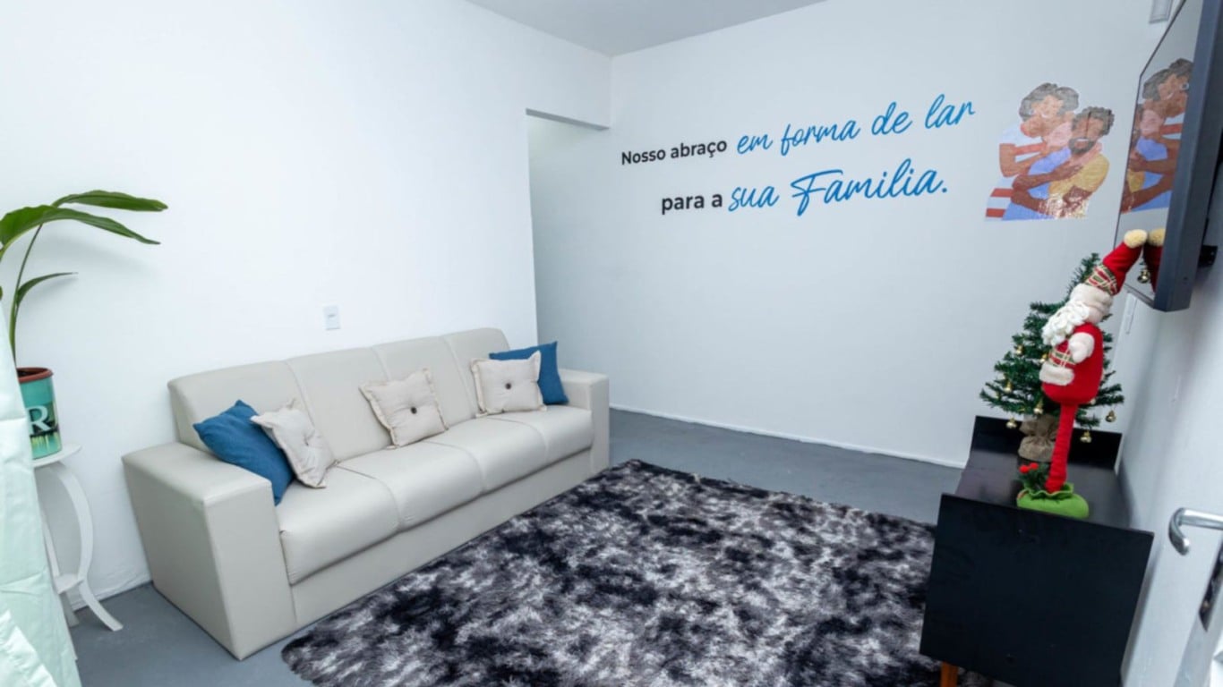 Sala da Casa da Família com sofá, tapete e frase “Nosso abraço em forma de lar para a sua família” na parede, além de decoração natalina. Sala da Casa da Família com sofá, tapete e frase “Nosso abraço em forma de lar para a sua família” na parede, além de decoração natalina.