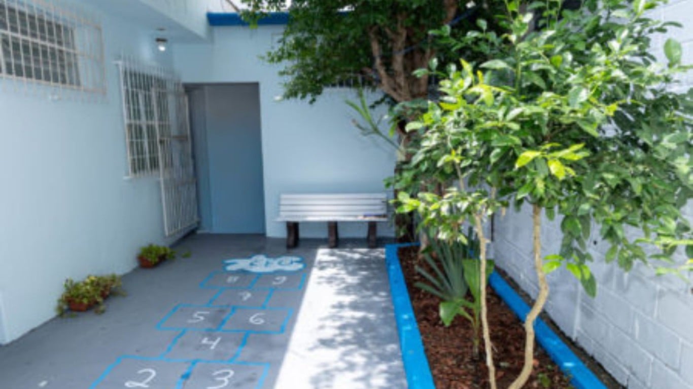 Entrada da Casa da Família com amarelinha pintada no chão, banco branco e árvores que compõem a área externa acolhedora. Entrada da Casa da Família com amarelinha pintada no chão, banco branco e árvores que compõem a área externa acolhedora.
