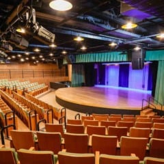 Interior da Sala Glória Rocha, no Centro das Artes de Jundiaí, com poltronas verdes, palco semicircular iluminado e equipamentos de iluminação no teto.