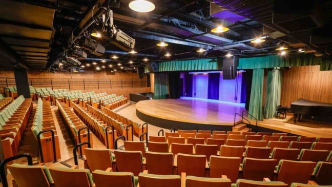 Interior da Sala Glória Rocha, no Centro das Artes de Jundiaí, com poltronas verdes, palco semicircular iluminado e equipamentos de iluminação no teto.