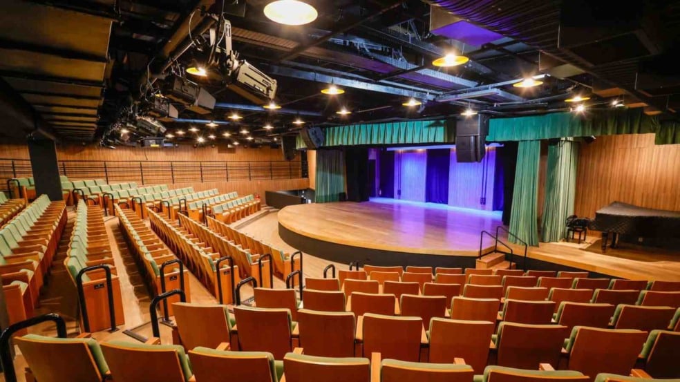 Interior da Sala Glória Rocha, no Centro das Artes de Jundiaí, com poltronas verdes, palco semicircular iluminado e equipamentos de iluminação no teto.