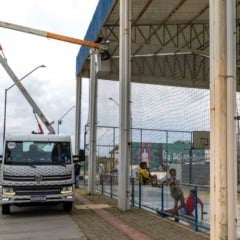 Caminhão com cesto elevatório instalado ao lado da quadra coberta do CEU das Artes Vista Alegre, onde técnicos trabalham na troca das lâmpadas, enquanto crianças jogam e observam o serviço.
