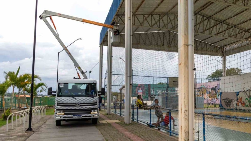 Caminhão com cesto elevatório instalado ao lado da quadra coberta do CEU das Artes Vista Alegre, onde técnicos trabalham na troca das lâmpadas, enquanto crianças jogam e observam o serviço. Caminhão com cesto elevatório instalado ao lado da quadra coberta do CEU das Artes Vista Alegre, onde técnicos trabalham na troca das lâmpadas, enquanto crianças jogam e observam o serviço.