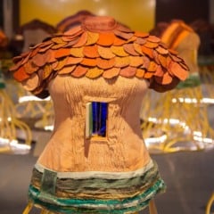 Escultura em forma de torso humano com placas têxteis coloridas nos ombros e uma pequena janela azul no centro, parte da instalação interativa Cidadela de Maria Ezou, com outras peças similares desfocadas ao fundo.