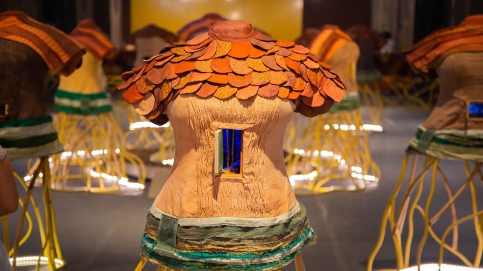 Escultura em forma de torso humano com placas têxteis coloridas nos ombros e uma pequena janela azul no centro, parte da instalação interativa Cidadela de Maria Ezou, com outras peças similares desfocadas ao fundo. Escultura em forma de torso humano com placas têxteis coloridas nos ombros e uma pequena janela azul no centro, parte da instalação interativa Cidadela de Maria Ezou, com outras peças similares desfocadas ao fundo.