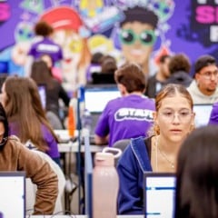 Jovens sentados em frente a computadores participam de um processo seletivo do CIEE, enquanto integrantes do staff, com camisetas roxas, auxiliam ao fundo em um ambiente movimentado e decorado com painel colorido.