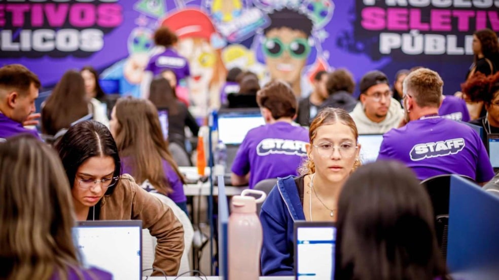Jovens sentados em frente a computadores participam de um processo seletivo do CIEE, enquanto integrantes do staff, com camisetas roxas, auxiliam ao fundo em um ambiente movimentado e decorado com painel colorido. Jovens sentados em frente a computadores participam de um processo seletivo do CIEE, enquanto integrantes do staff, com camisetas roxas, auxiliam ao fundo em um ambiente movimentado e decorado com painel colorido.