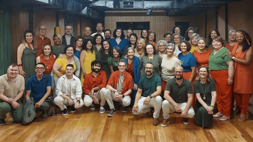 Integrantes da Cia. Canto Vivo posam juntos para foto em um estúdio de ensaio, organizados em três fileiras e sorrindo. O grupo é diverso e veste roupas coloridas em tons vibrantes, transmitindo unidade e alegria.
