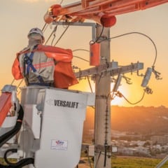 Eletricista em plataforma elevatória realizando manutenção na rede elétrica ao pôr do sol, com vista da cidade ao fundo.