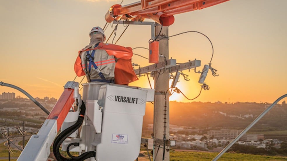 Eletricista em plataforma elevatória realizando manutenção na rede elétrica ao pôr do sol, com vista da cidade ao fundo. Eletricista em plataforma elevatória realizando manutenção na rede elétrica ao pôr do sol, com vista da cidade ao fundo.