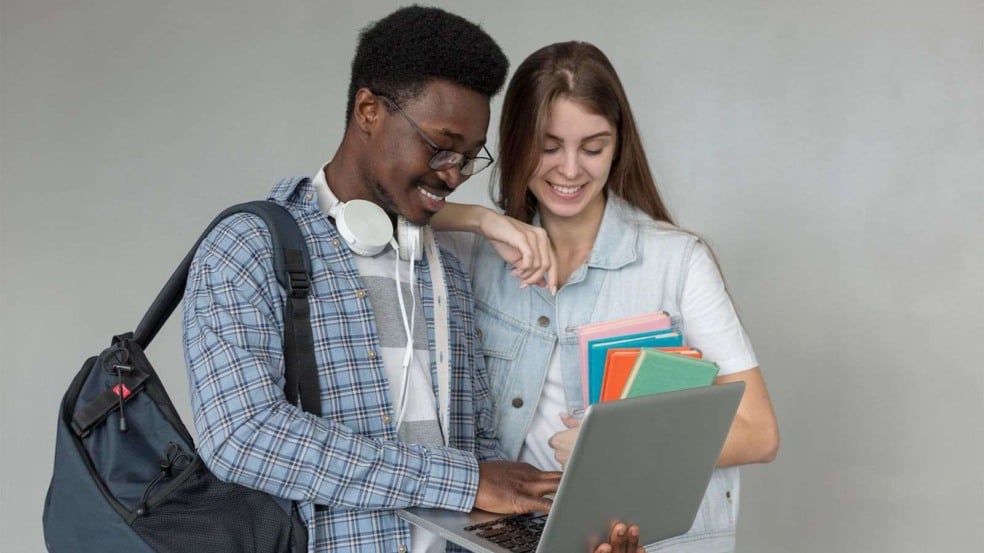 Jovem estudante sorrindo enquanto digita em um notebook ao lado de uma colega que segura cadernos e observa a tela. Jovem estudante sorrindo enquanto digita em um notebook ao lado de uma colega que segura cadernos e observa a tela.
