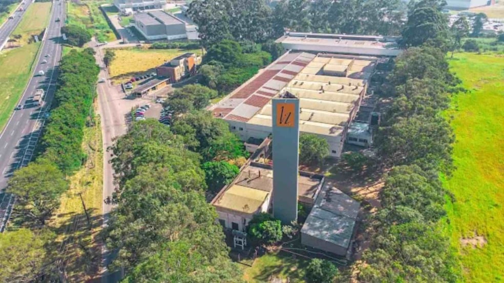 Vista aérea de um complexo industrial com galpões grandes, torre com logotipo laranja e áreas verdes ao redor, ao lado da Rodovia Anhanguera em Jundiaí.